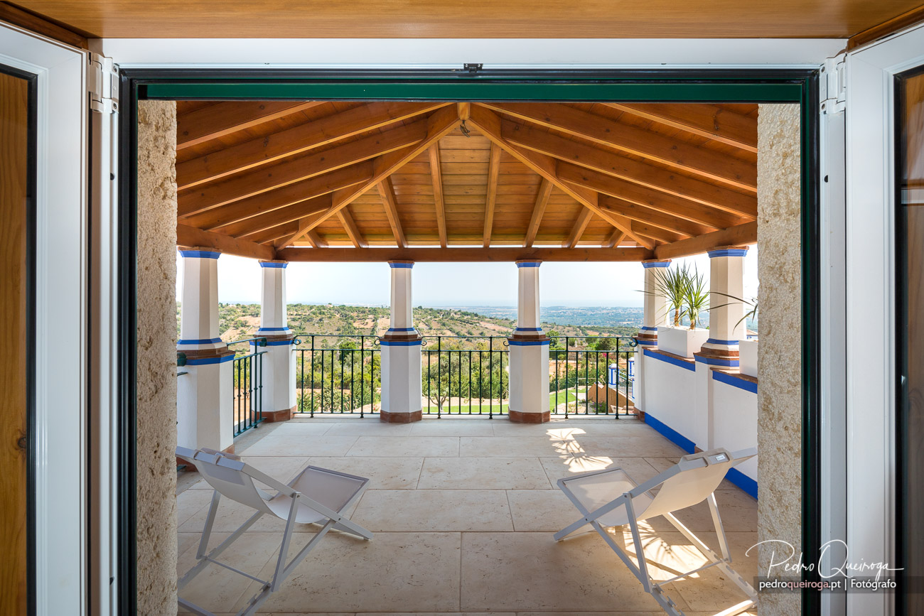 Varanda ampla com telhado de madeira, colunas brancas e azuis, duas espreguiçadeiras e vista panorâmica para o campo alentejano.