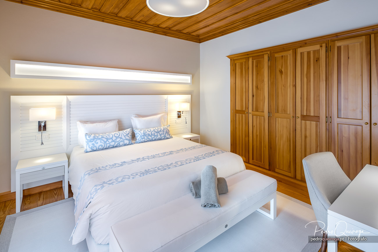 Quarto amplo com cama branca e roupa azul, roupeiros em madeira, teto em lambrim e iluminação suave e acolhedora.