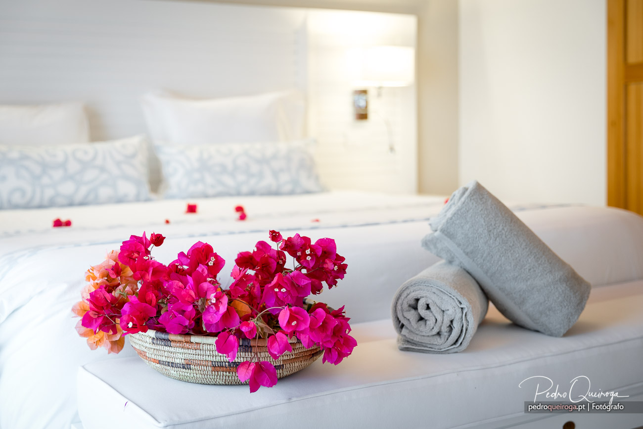 Quarto de hotel luminoso com flores cor-de-rosa vibrantes em cesto e toalhas cinzentas enroladas sobre a cama branca.