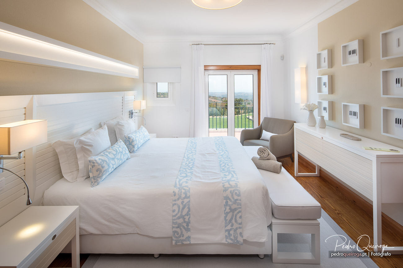 Quarto luminoso com tons claros, molduras fotográficas na parede e vista panorâmica sobre a paisagem verde