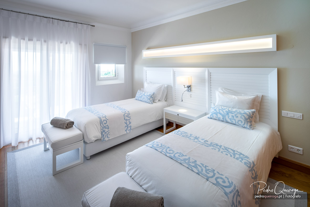 Quarto duplo com arquitetura de interiores clara, iluminação LED e têxteis em branco e azul suave.