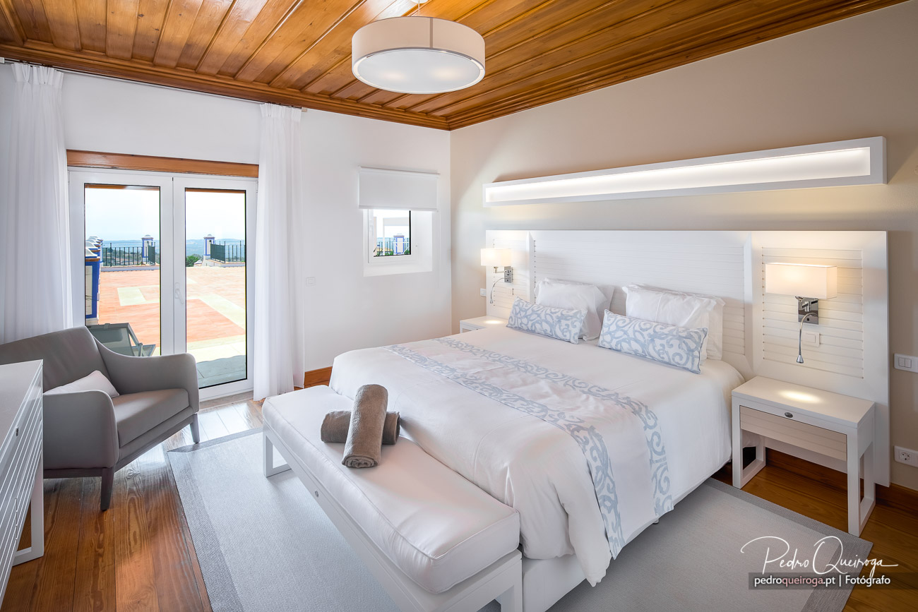 Quarto moderno com teto em madeira, cama de casal branca, poltrona cinzenta e acesso a terraço com vista panorâmica