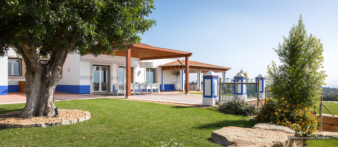 Moradia moderna com pérgola de madeira, relvado verdejante, árvore frondosa e detalhes azuis na fachada branca.