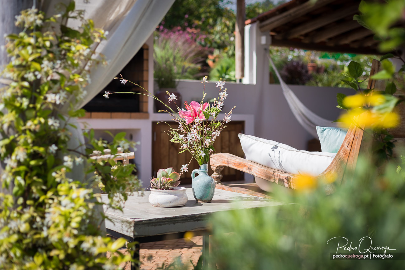 Terraço acolhedor com espreguiçadeira de madeira, mesa com flores em jarro azul e vegetação verdejante em redor