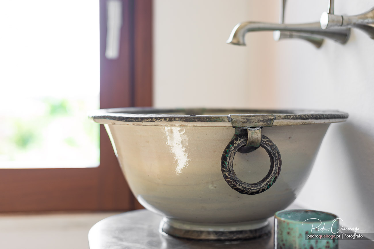 Bacia vintage em cerâmica bege com asa de argola em metal oxidado sobre base branca num ambiente luminoso