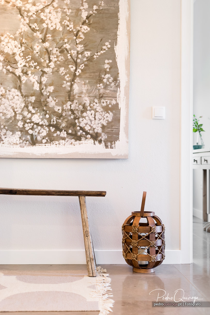 Quadro decorativo com flores brancas sobre parede clara, banco de madeira e lanterna âmbar em interior luminoso.