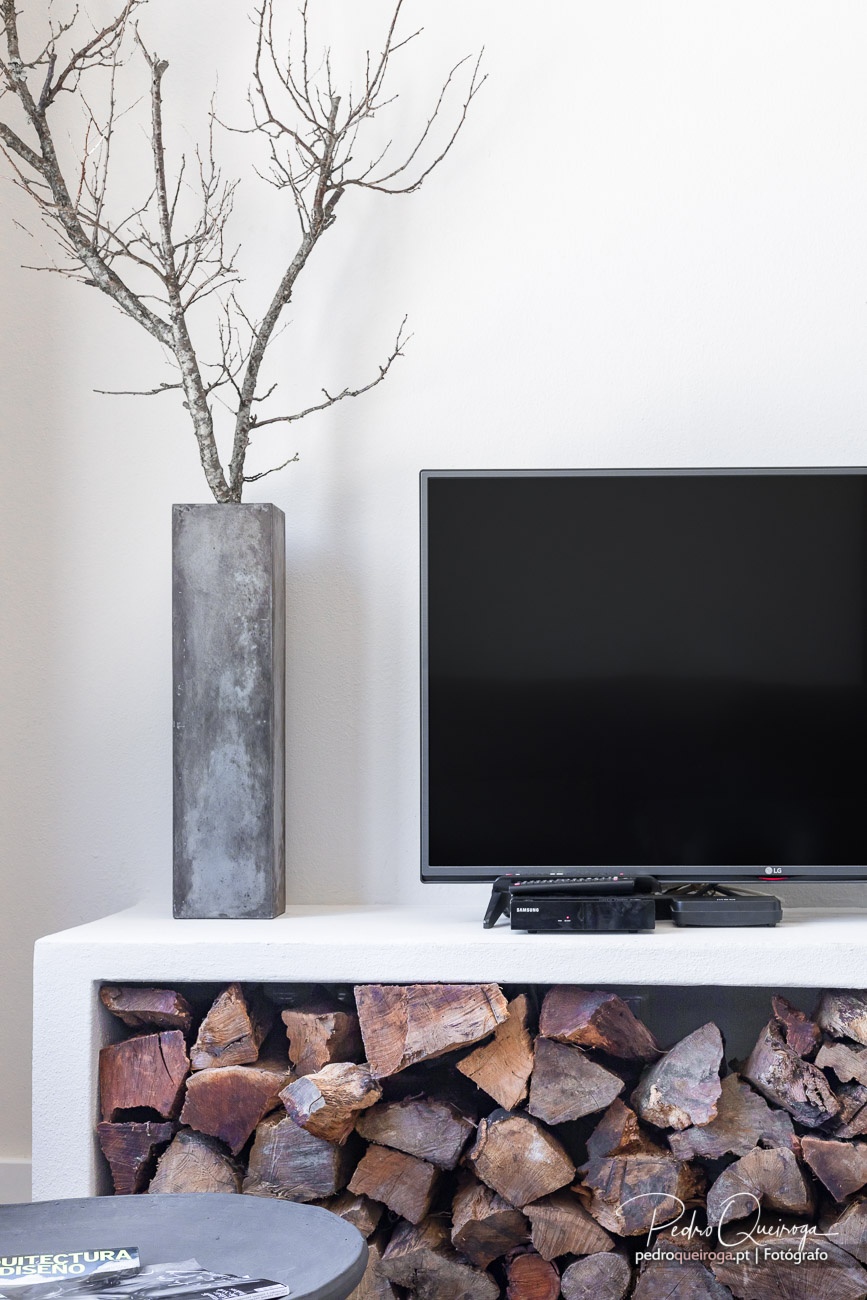 Sala minimalista com televisão sobre móvel branco, vaso cinzento com ramos secos e lenha arrumada por baixo.