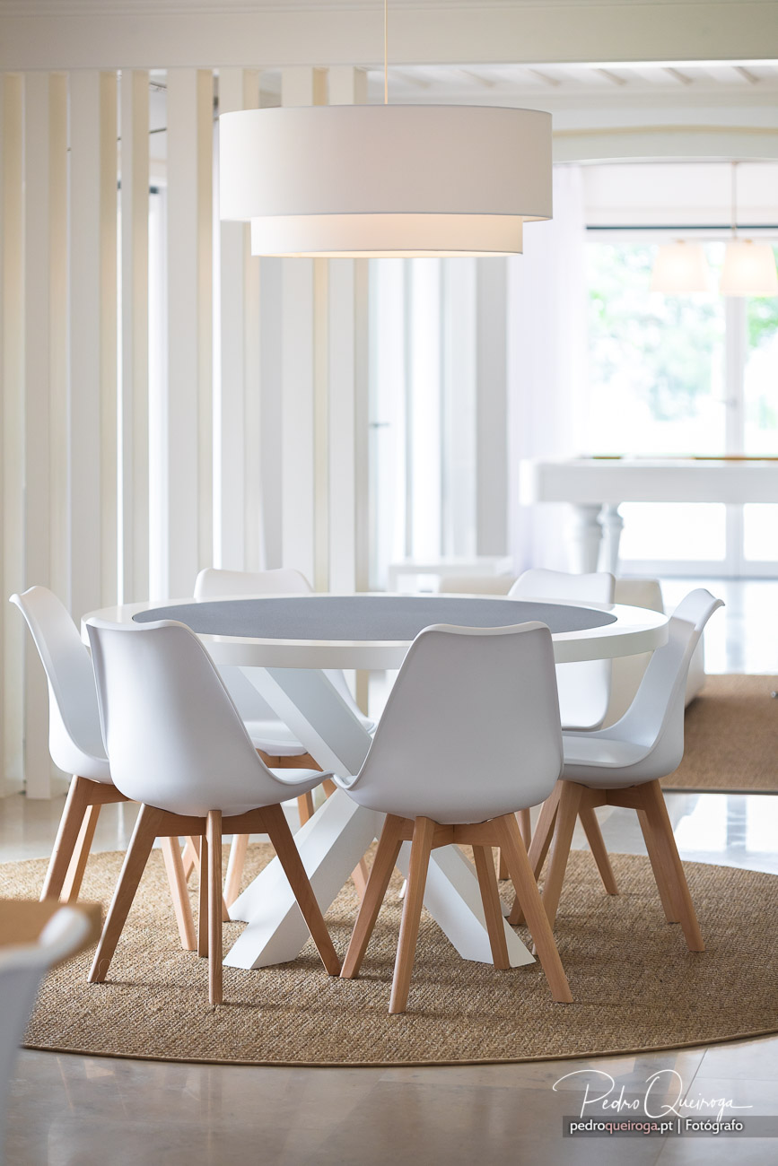 Sala de jantar moderna com mesa redonda cinzenta, cadeiras brancas e candeeiro suspenso em tons claros e luminosos