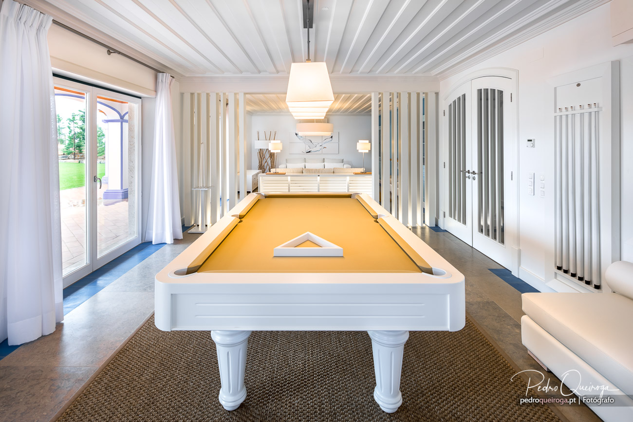 Sala de jogos luminosa com mesa de bilhar branca e feltro amarelo, divisórias verticais e teto em ripado de madeira