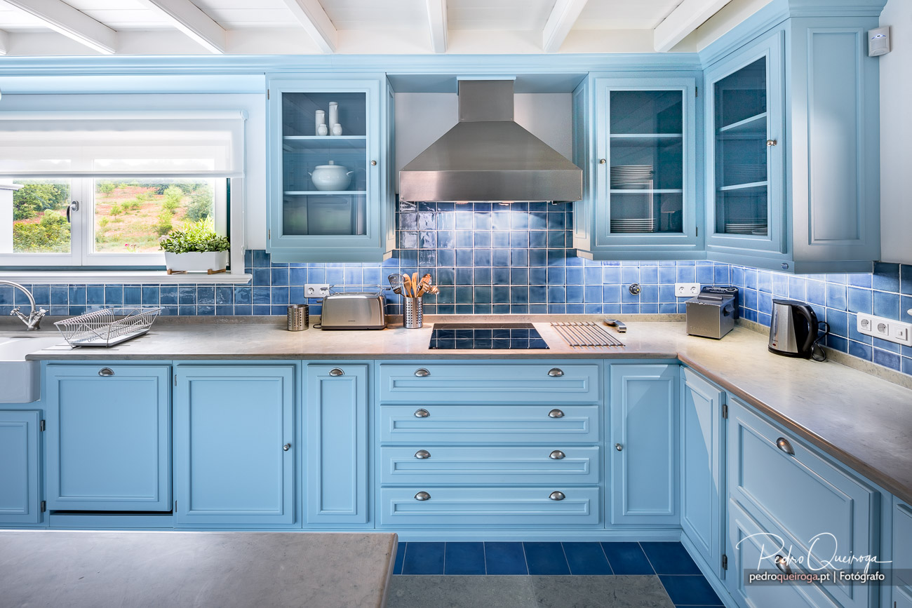 Cozinha moderna em tons azul pastel com azulejos brilhantes, bancada cinza, exaustor inox e janela ampla com vista natural.