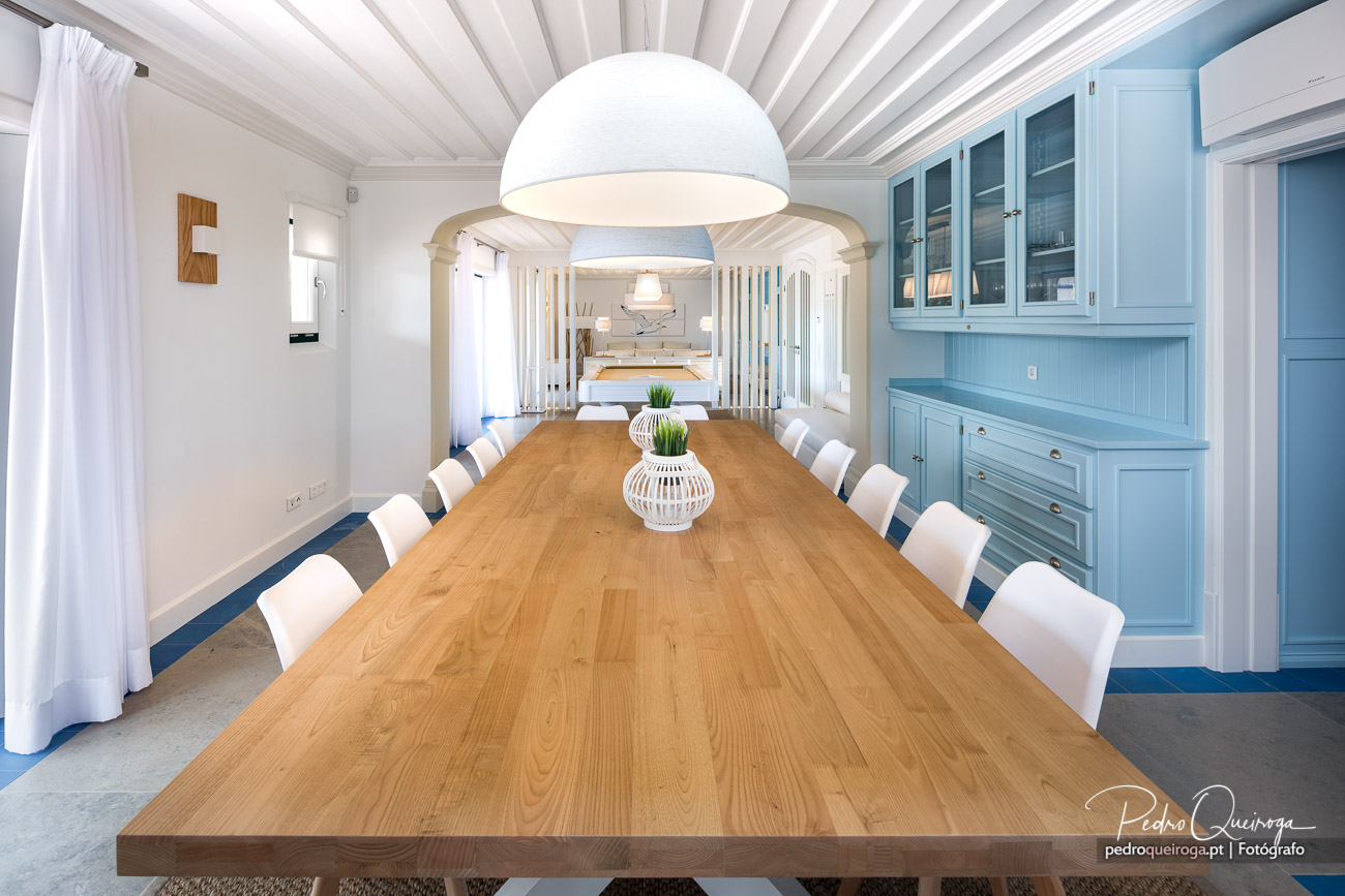 Sala de jantar luminosa com mesa em madeira, cadeiras brancas e móvel azul claro, teto em madeira branca