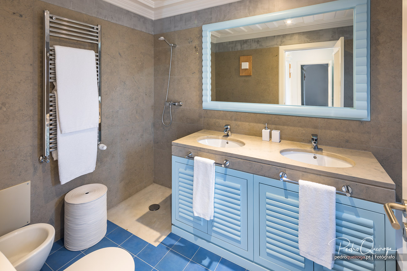 Casa de banho moderna com móvel azul de duplo lavatório, espelho iluminado, toalheiro aquecido e chuveiro em tons bege.