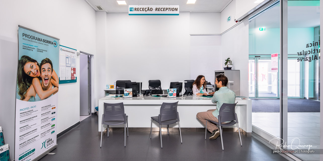 Balcão de receção moderno com funcionária a atender cliente sentado, paredes brancas e sinalética no teto