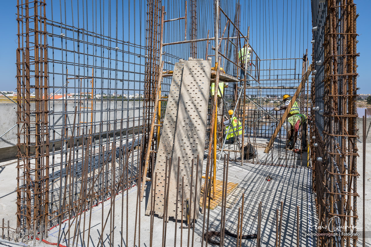 Operários em andaime a trabalhar em armaduras de aço para pilares de betão armado em obra sob céu azul