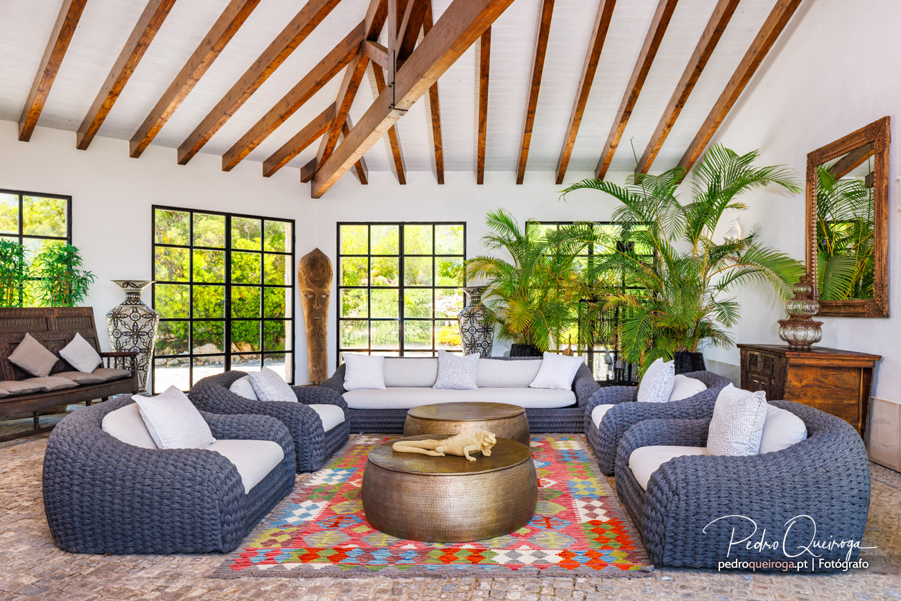 Sala ampla com tecto de vigas de madeira, sofás em fibra natural, plantas tropicais e janelas com vista para jardim