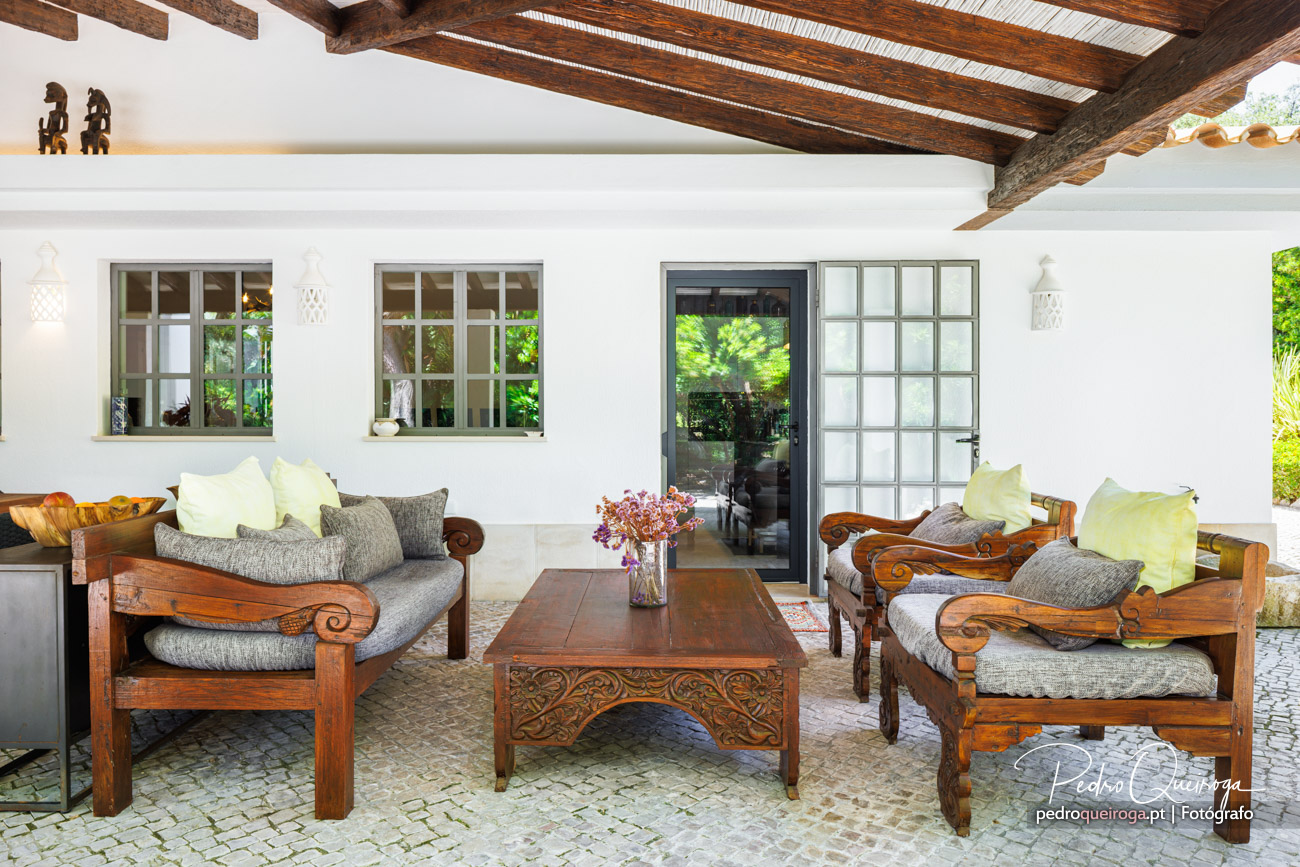 Alpendre elegante com sofás de madeira trabalhada, almofadas claras, mesa de centro e teto com vigas à vista rústicas.