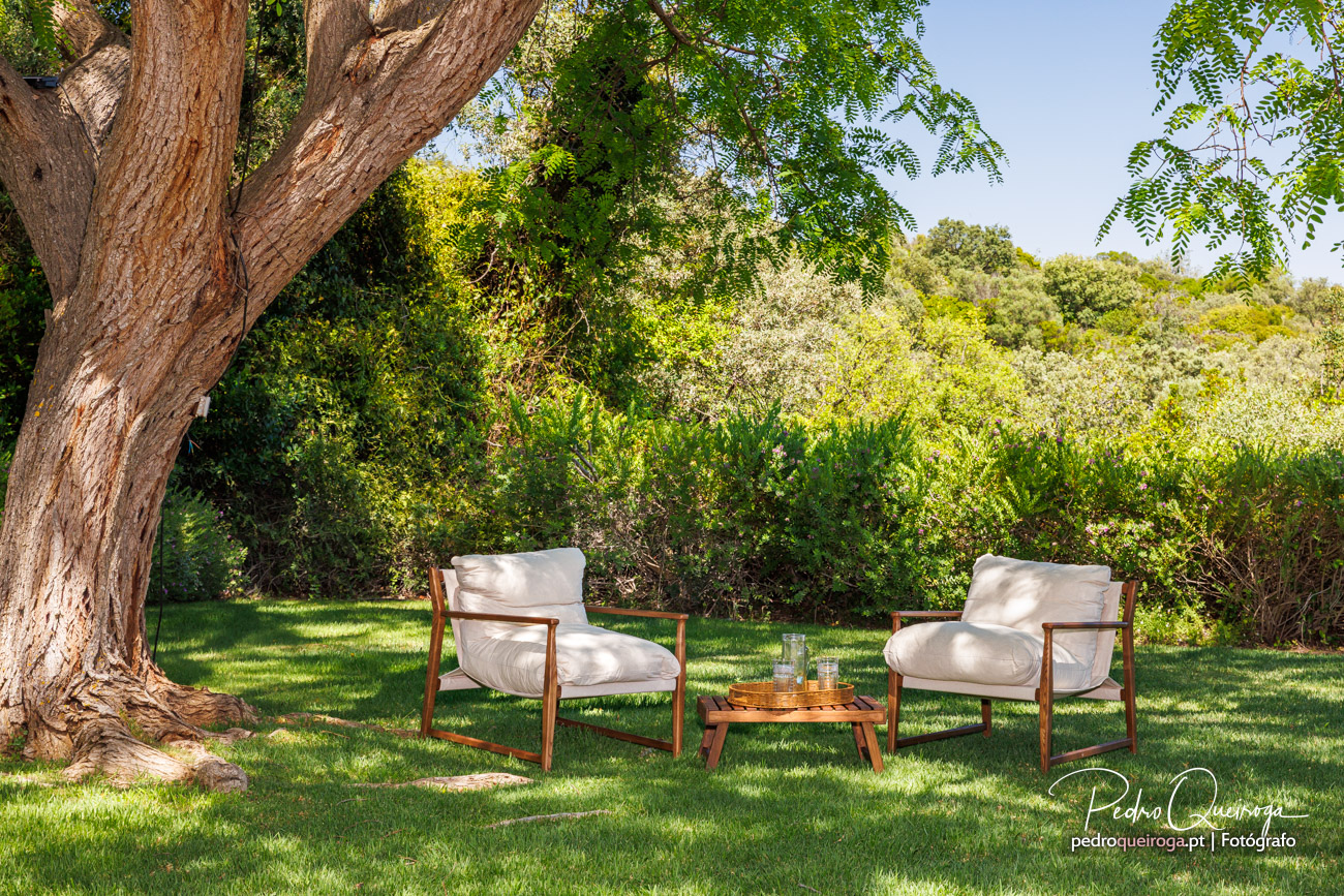Duas cadeiras brancas com estrutura em madeira e mesa baixa num jardim verdejante sob árvore frondosa em dia ensolarado.