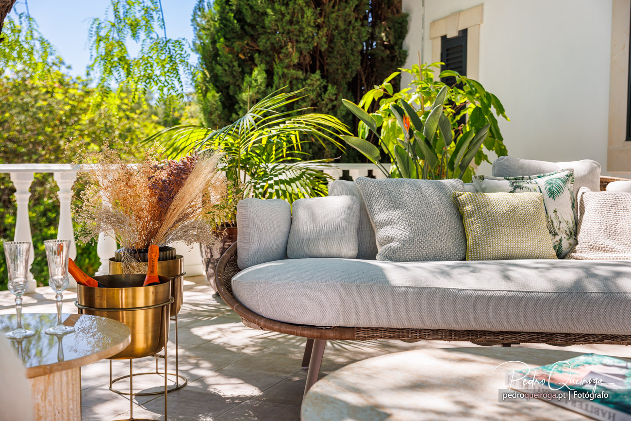 Terraço ensolarado com sofá em tons cinza e almofadas decorativas, rodeado por vegetação tropical e detalhes dourados elegantes.