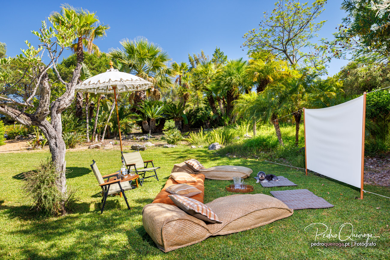 Cinema ao ar livre num jardim tropical com espreguiçadeiras, guarda-sol balinês e vegetação exuberante ao fundo