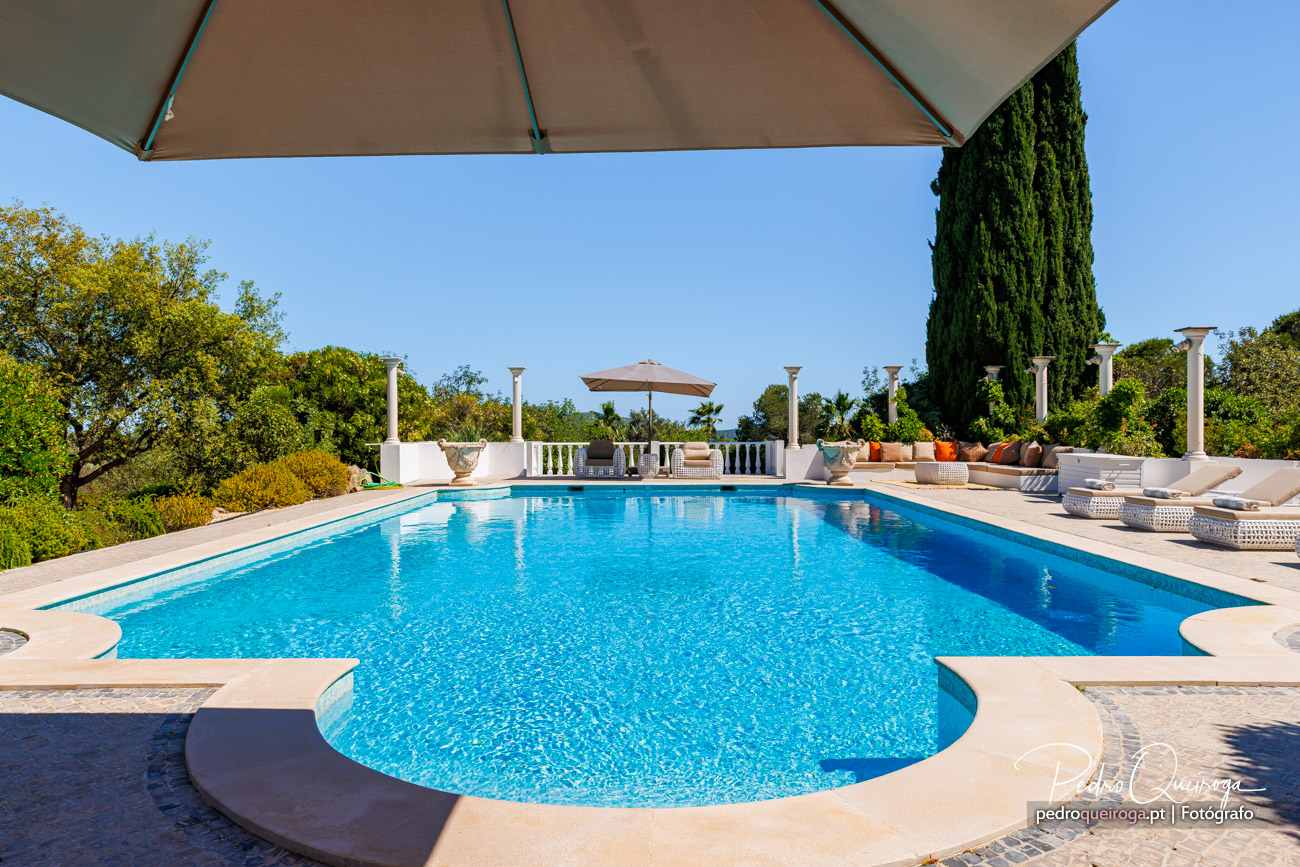 Piscina de luxo com colunas brancas, chapéus de sol bege e zona lounge rodeada por ciprestes e céu azul