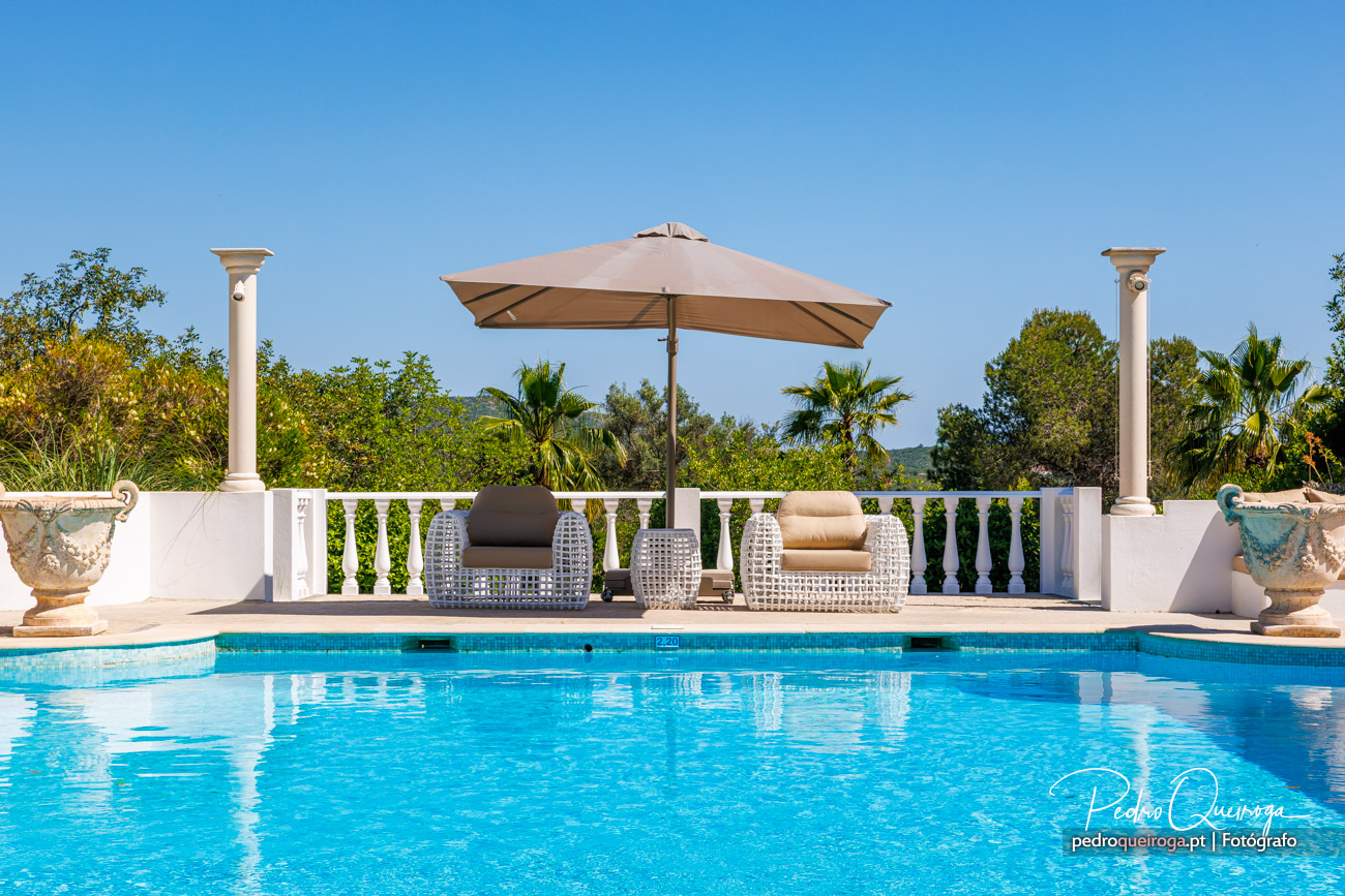Piscina azul cristalina com zona lounge, chapéu-de-sol bege, colunas brancas e vegetação tropical ao fundo sob céu azul.