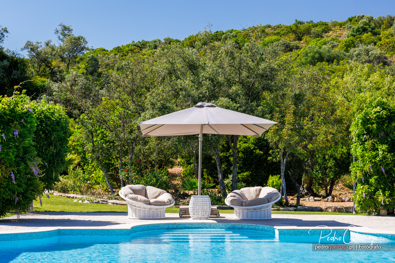 Piscina exterior de hotel com espreguiçadeiras em vime, chapéu de sol bege e paisagem natural verdejante ao fundo