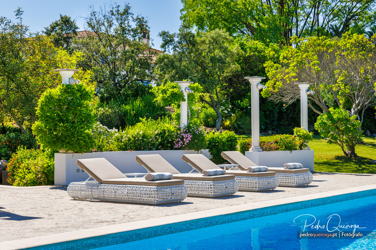 Espreguiçadeiras elegantes junto à piscina com colunas brancas e decoração de jardim exuberante ao fundo