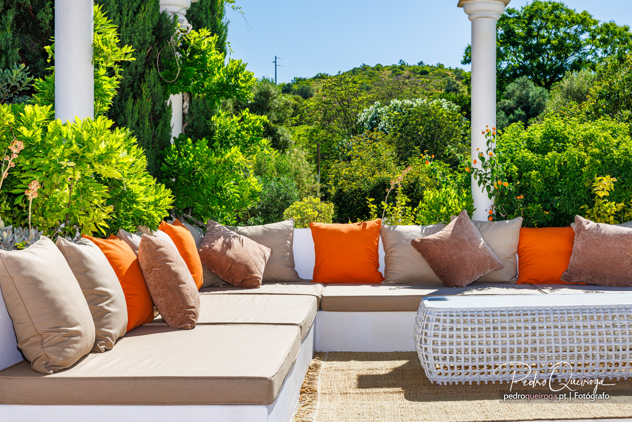 Terraço elegante com sofás bege e almofadas laranja, rodeado por vegetação exuberante e colunas brancas sob céu azul.