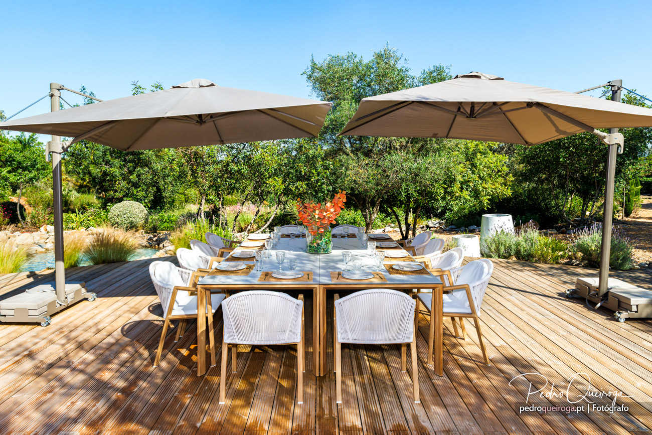 Mesa de exterior em madeira com cadeiras brancas sob chapéus de sol bege num deck rodeado por jardim mediterrânico