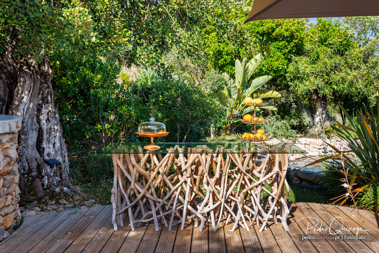 Mesa rústica de madeira em jardim exuberante, com bananeiras e árvores ao fundo, sob luz natural suave.