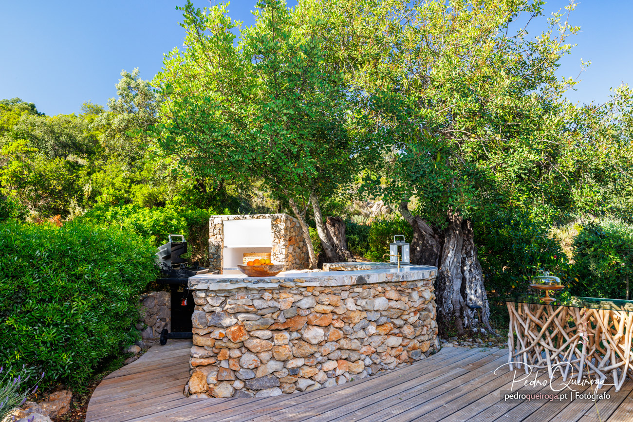 Barbecue exterior em pedra rústica sob árvore frondosa, rodeado por deck de madeira e vegetação exuberante num dia ensolarado