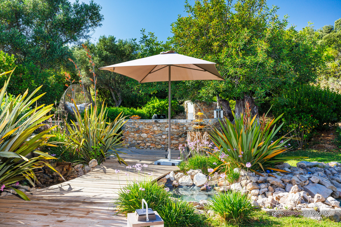 Jardim ensolarado com deck de madeira, chapéu-de-sol bege, plantas tropicais e muro em pedra ao fundo rodeado de árvores