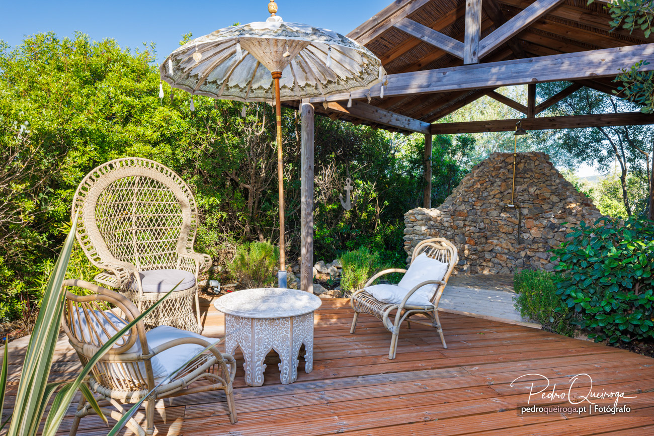 Terraço em madeira com mobiliário em vime, guarda-sol branco e pérgola junto a parede em pedra rodeada de vegetação