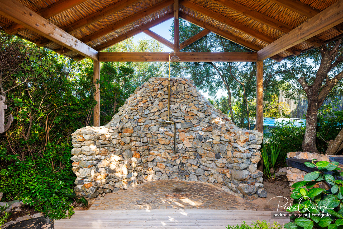 Duche exterior em pedra natural sob pérgola de caniço, rodeado de vegetação mediterrânica no Algarve