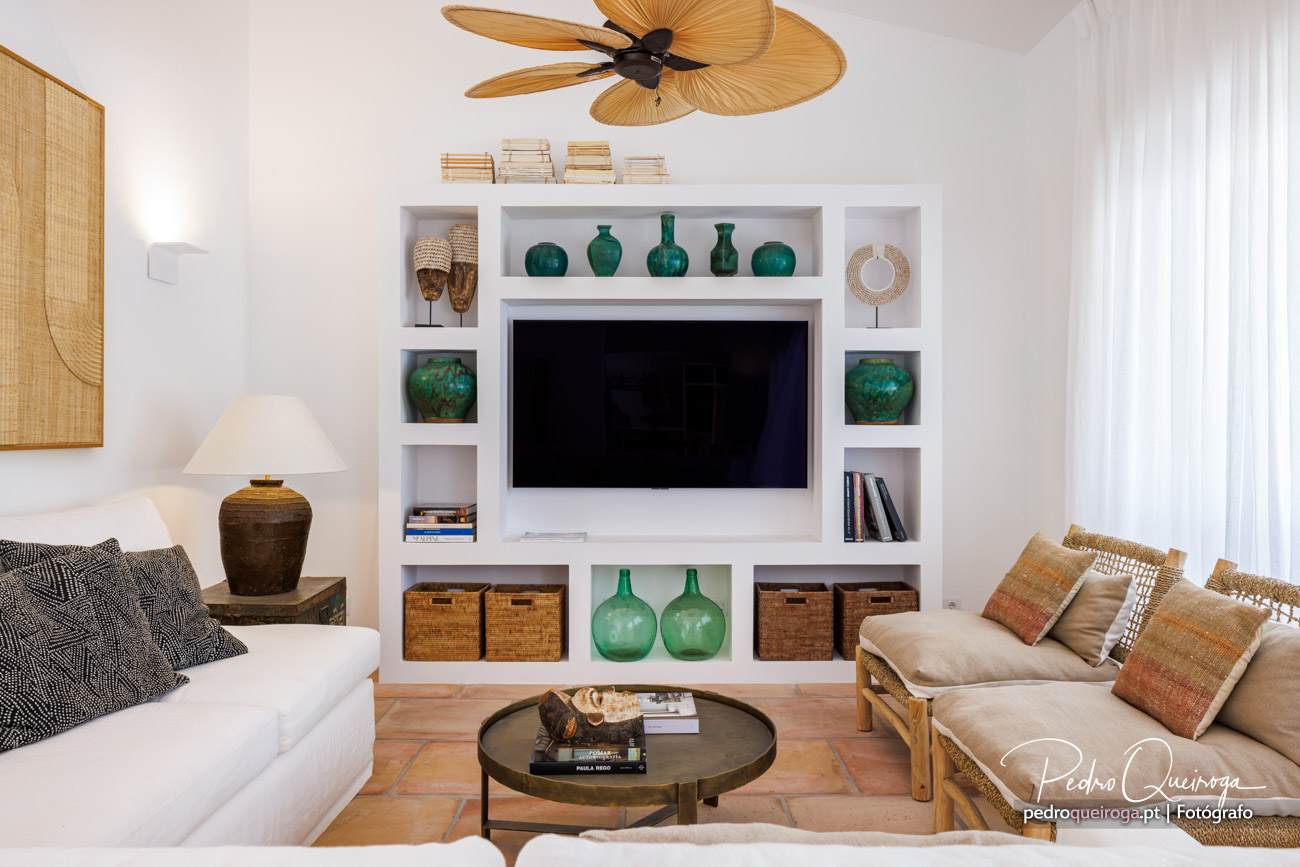 Sala de estar luminosa com estante branca, televisão integrada, peças decorativas verdes e mobiliário natural em tons bege.