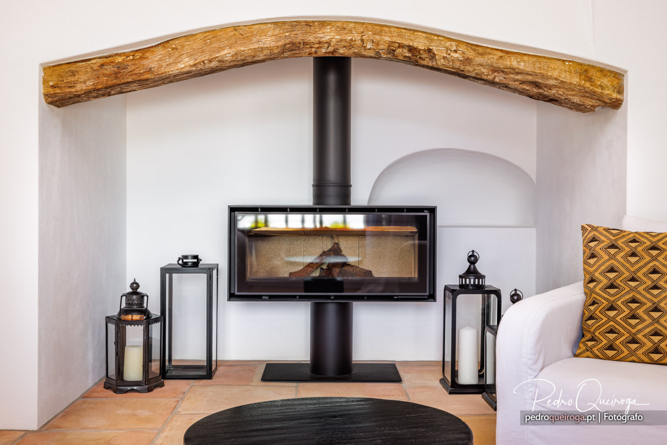 Salamandra moderna preta com porta de vidro sob viga de madeira rústica, ladeada por lanternas decorativas em sala branca