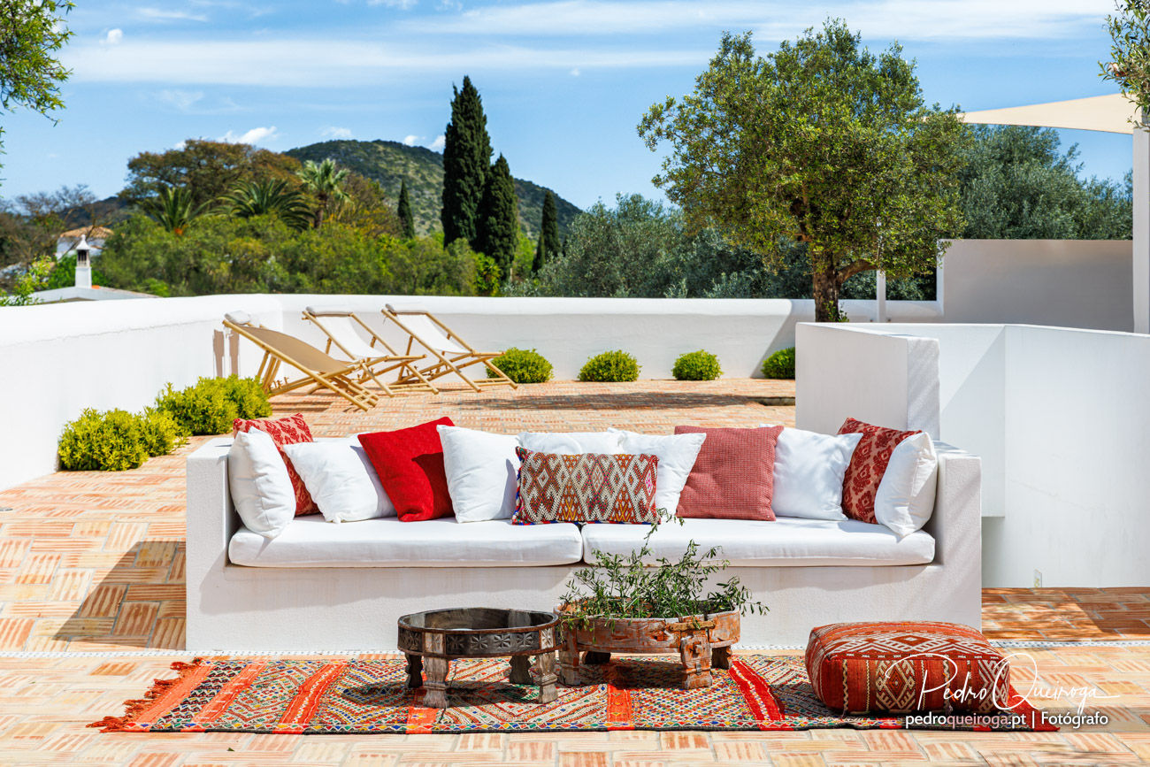 Terraço moderno com sofá branco, almofadas coloridas e espreguiçadeiras, rodeado por vegetação e montanhas ao fundo.