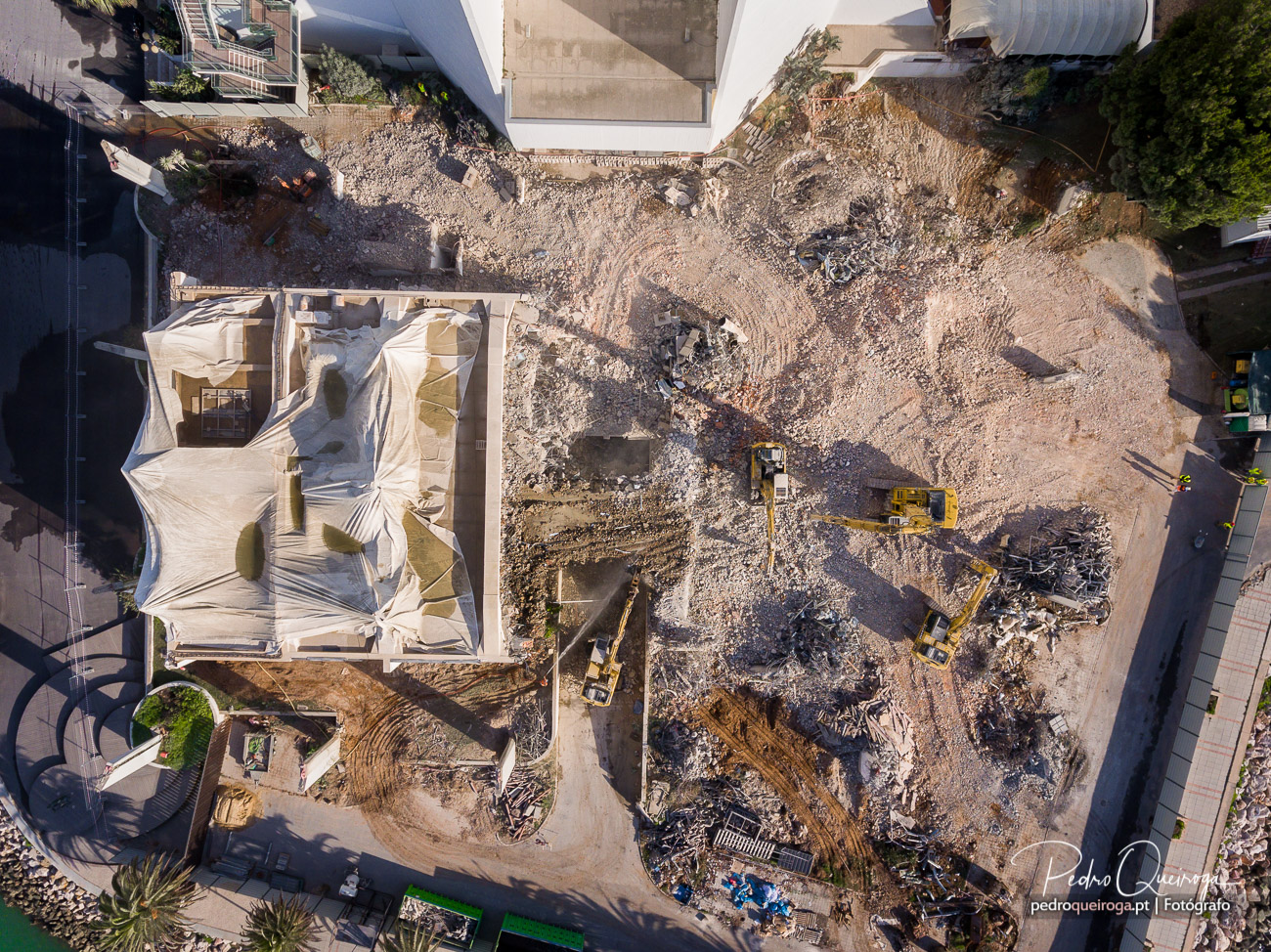 Vista aérea zenital do estaleiro de demolição com estrutura coberta por lona e escavadoras em operação