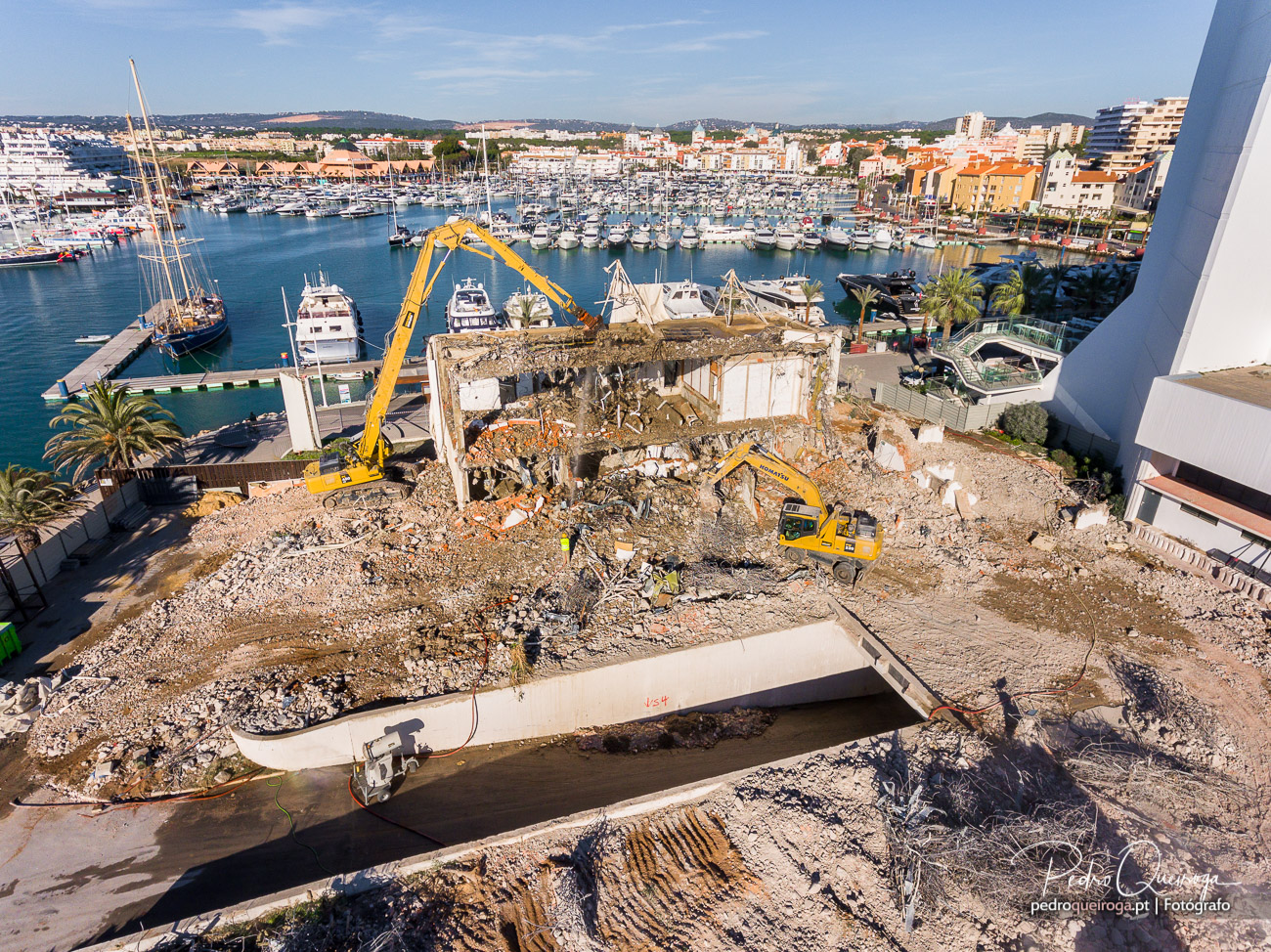 Vista aérea lateral da demolição com escavadoras de grande alcance e marina de Vilamoura ao fundo