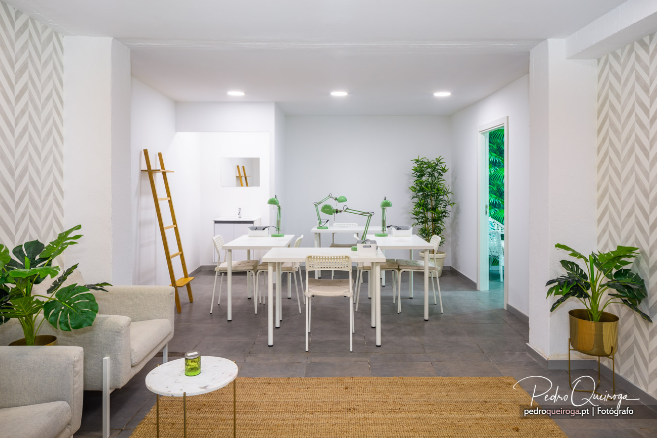 Espaço de trabalho amplo e luminoso com mesas brancas, candeeiros verdes e plantas decorativas em tons naturais