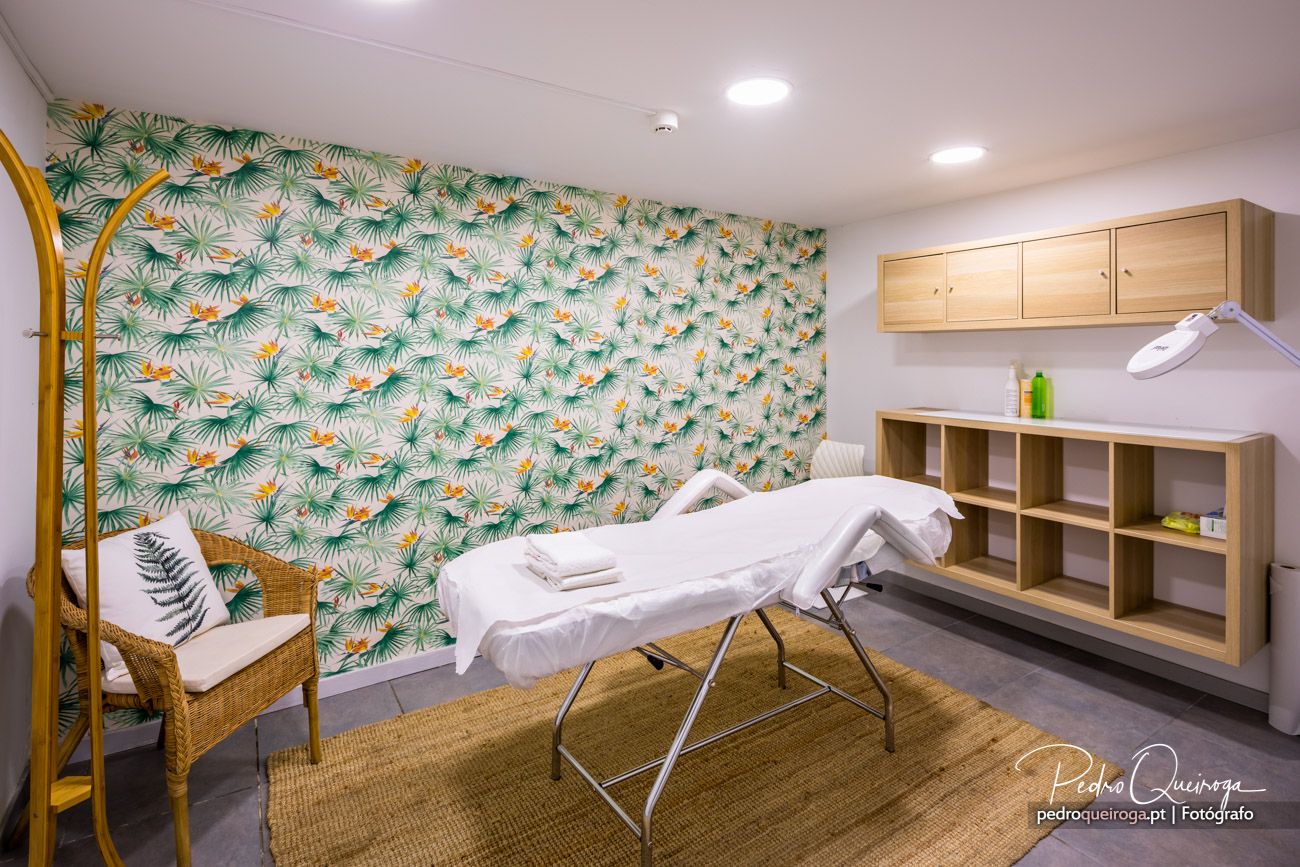 Sala de massagens com papel de parede tropical, maca branca, móveis em madeira clara e cadeira de vime sobre tapete natural.