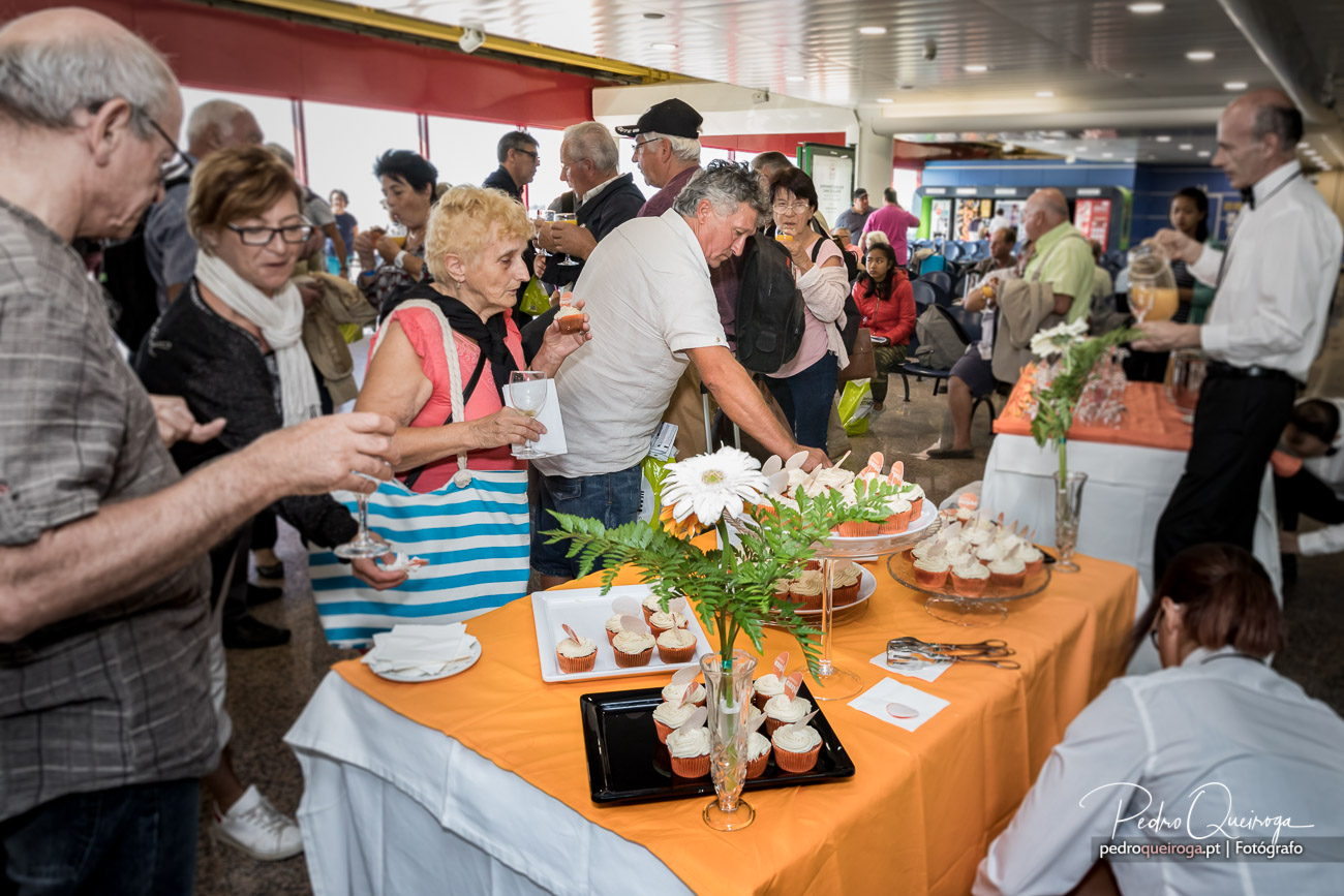 Grupo de pessoas reunido junto a mesa com toalha laranja repleta de cupcakes e flores durante evento social animado