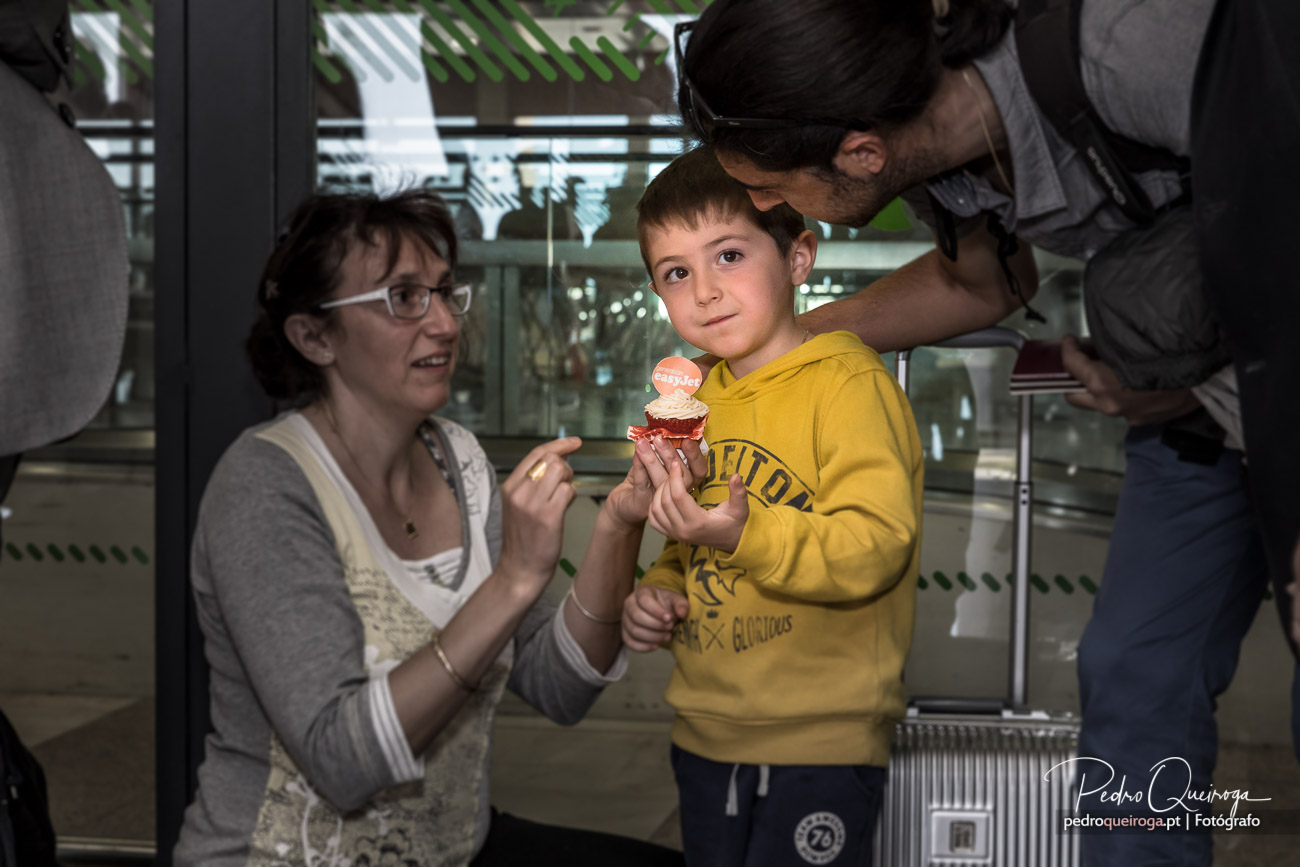 Criança de camisola amarela segura cupcake junto aos pais numa pastelaria, observando com curiosidade e sorriso tímido.