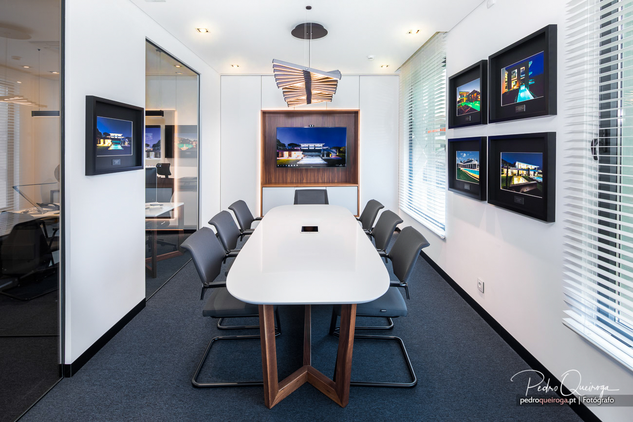 Sala de reuniões moderna com mesa branca, cadeiras cinzentas, ecrã embutido e fotografias emolduradas nas paredes
