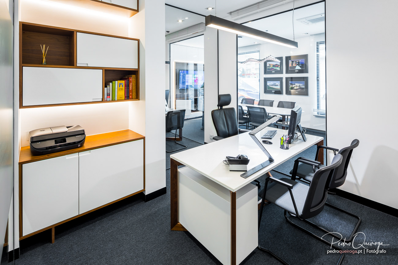 Escritório moderno com mobiliário branco e madeira, decorado com fotografias emolduradas na sala de reuniões ao fundo.