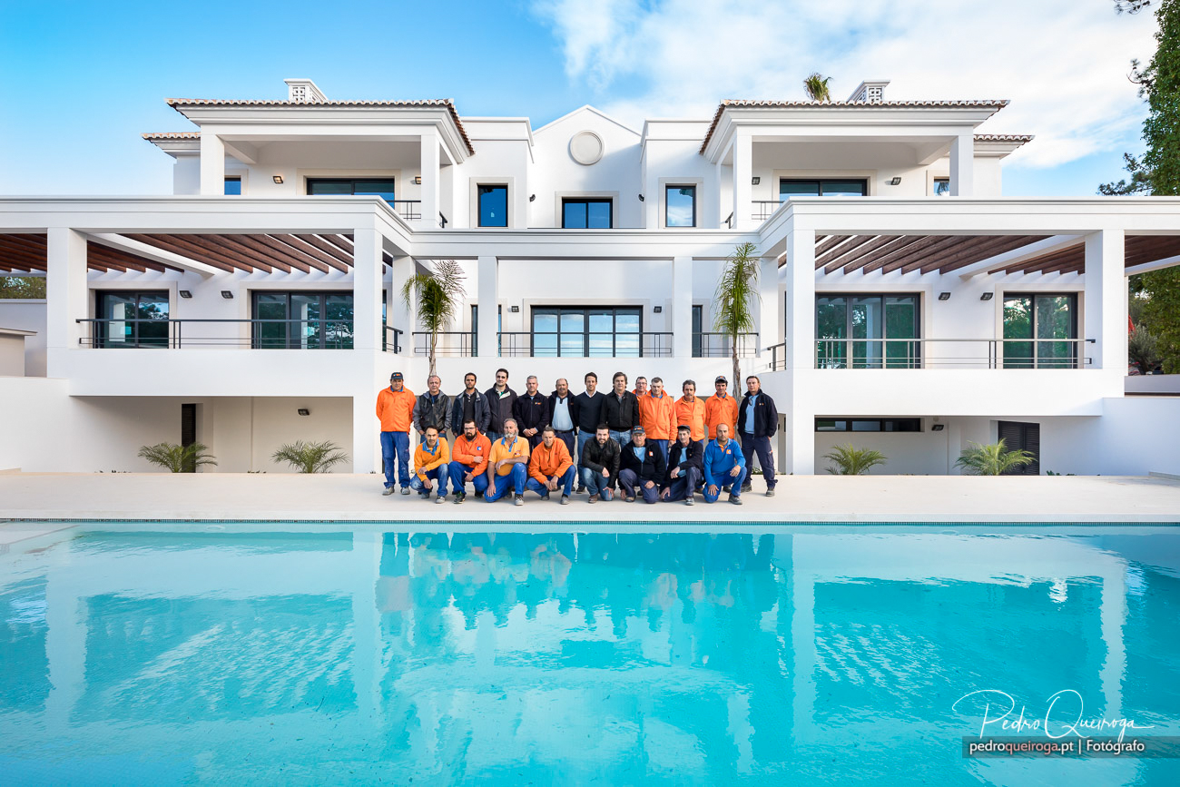 Equipa de construção posa em frente a moradia branca moderna com piscina sob céu azul e nuvens dispersas