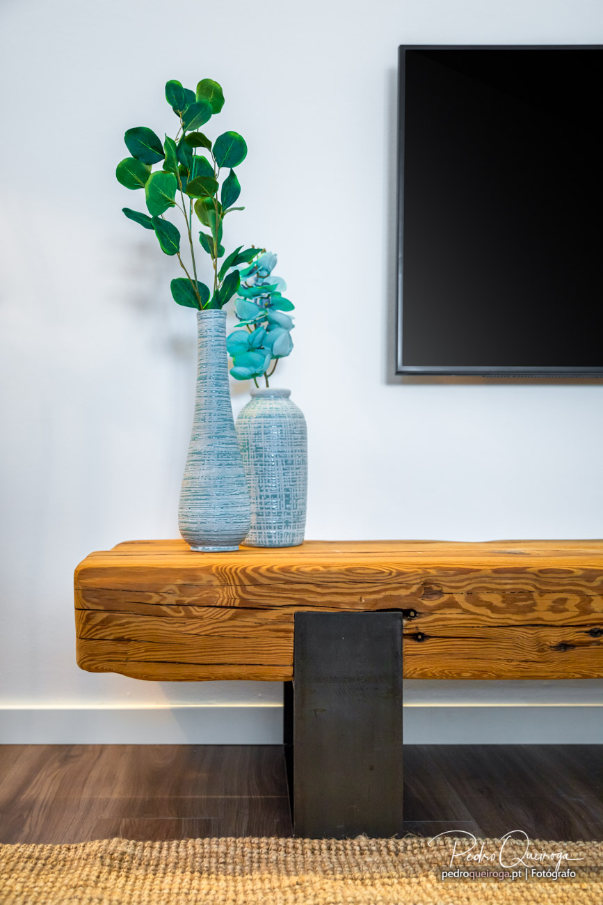 Detalhe decorativo de imóvel com móvel em madeira, vasos azuis turquesa e ramos verdes sob quadro moderno na parede