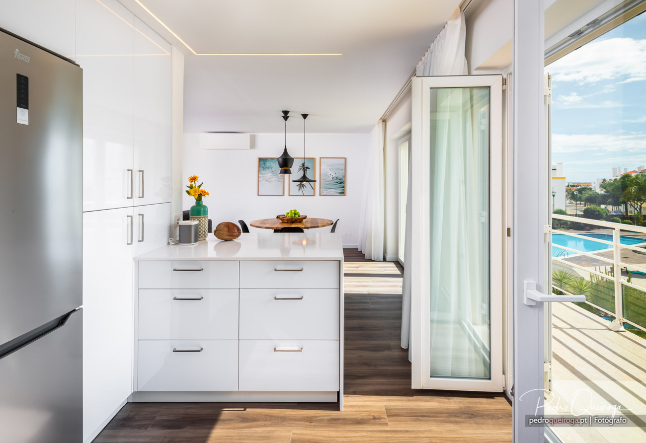 Cozinha moderna e luminosa com móveis brancos, quadros na parede e varanda com vista para piscina e jardim.