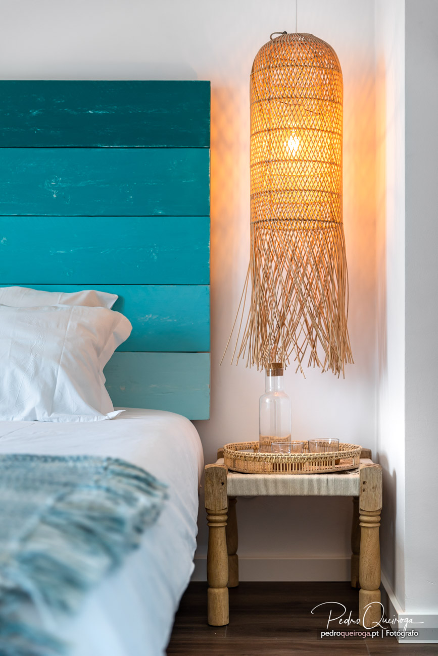 Quarto acolhedor com cabeceira turquesa, candeeiro de vime artesanal aceso e mesinha de apoio em madeira natural