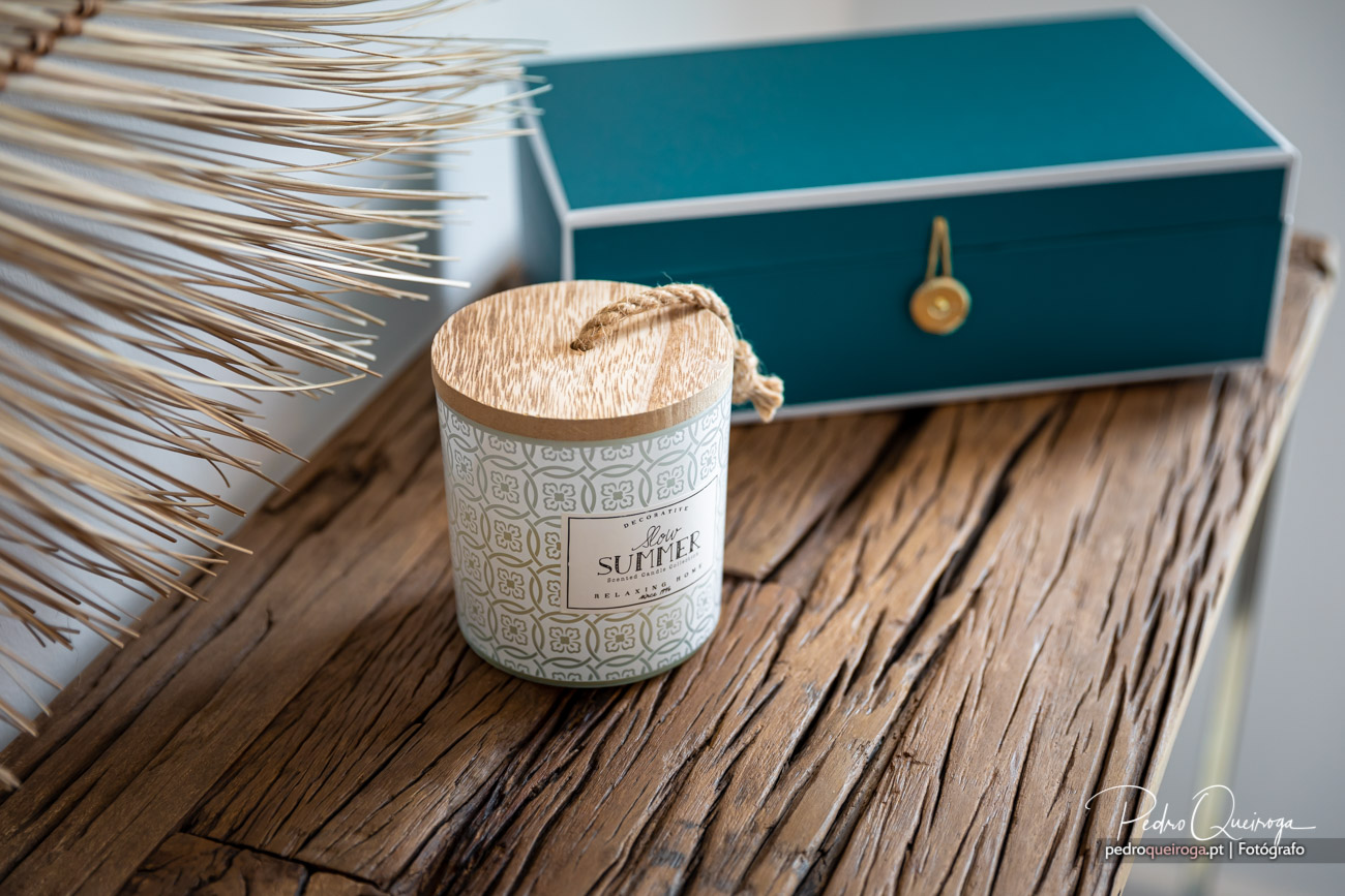 Vela aromática com tampa de madeira sobre mesa rústica, ao lado de caixa azul turquesa e folhas decorativas secas.